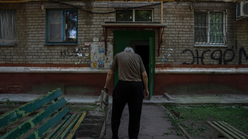 An old man walks away from camera toward a building covered in graffiti and with windows broken. 