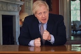 British PM Boris Johnson sits at a table in a dark suit with fists clenched.