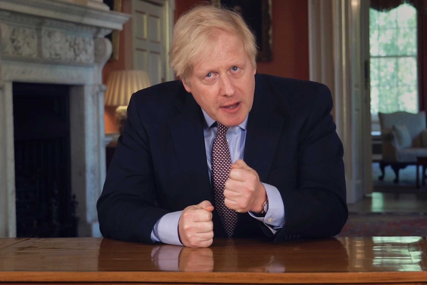 British PM Boris Johnson sits at a table in a dark suit with fists clenched.