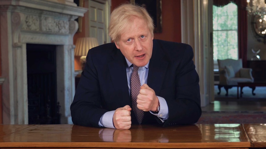 British PM Boris Johnson sits at a table in a dark suit with fists clenched.