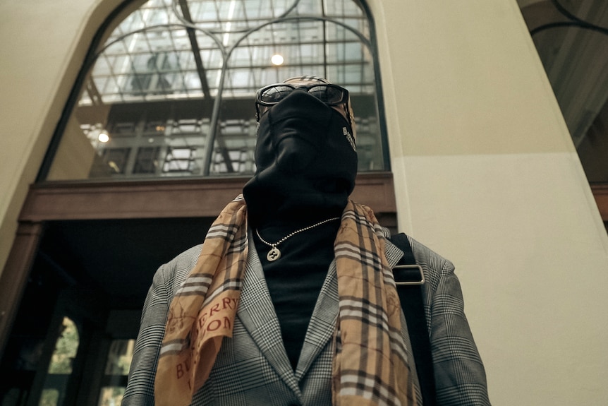 A woman in corporate attire with a scarf, mask and glasses covering her face looks up while standing in an ornate building.