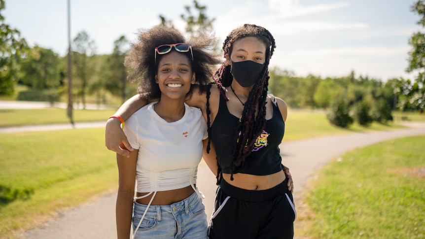 Two young women in a park, one wearing a mask with her arm around her friend's shoulders. Both smile brightly