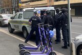 Police gather around the back of a wagon in a city street with purple scooters in it