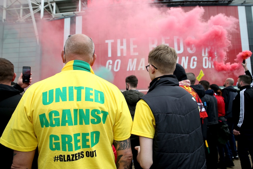 A fan wearing a green and yellow scarf and a shirt with 'United Against Greed' on it, faces red smoke from flares.