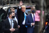 Donald Trump standing in the street near a car waves while surrounded by men in suits with surgical masks on.