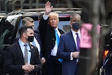 Donald Trump standing in the street near a car waves while surrounded by men in suits with surgical masks on.