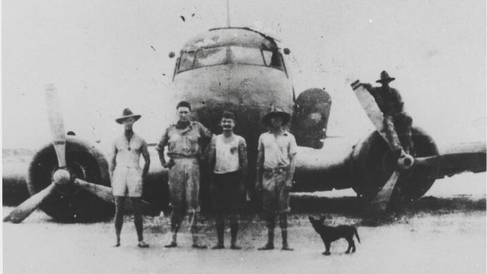 Black and white photo of four men and a dog standing in front of a plane wreck in the sand. Another man is on a propeller.