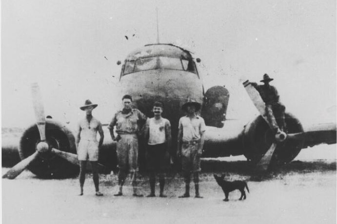 Black and white photo of four men and a dog standing in front of a plane wreck in the sand. Another man is on a propeller.