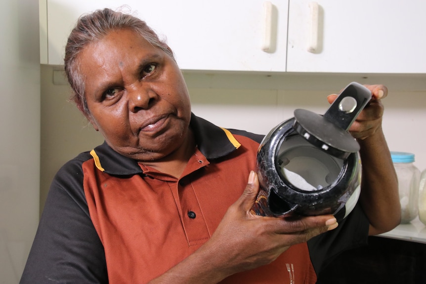 A woman is in her kitchen holding up a kettle. Her expression is serious.