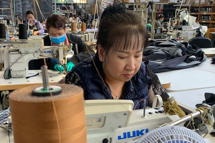 A woman sits over a sewing machine making jeans in a factory with other workers bent over sewing machines behind her.