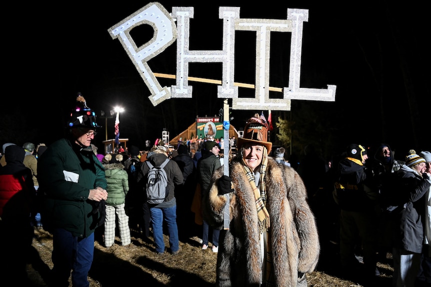 A woman in winter fur coat holds a sign of cutout letters reading P H I L 