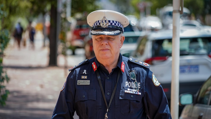 A man in a NT Police uniform walking along a city footpath and looking serious, on a sunny day.