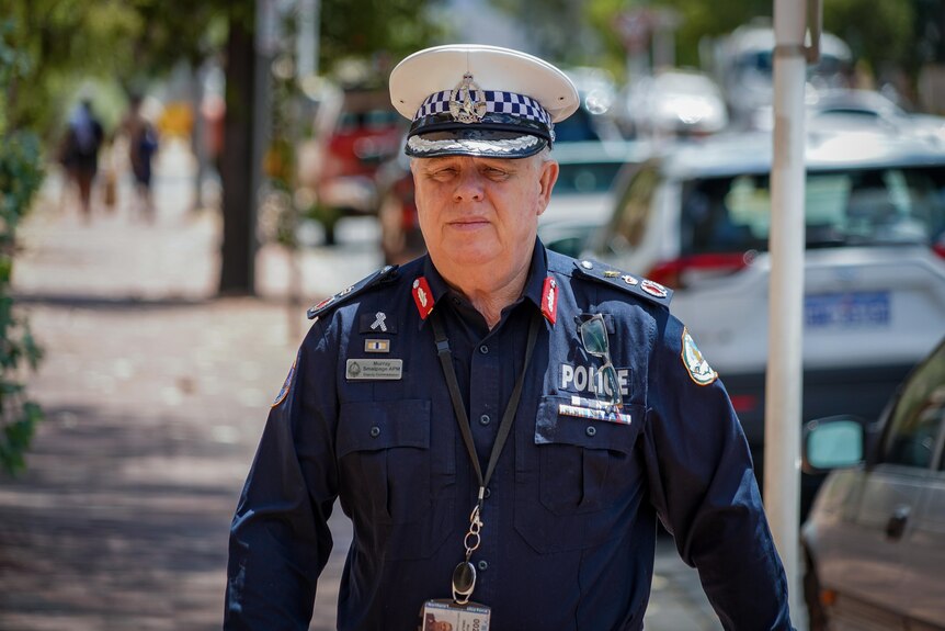 A man in a NT Police uniform walking along a city footpath and looking serious, on a sunny day.