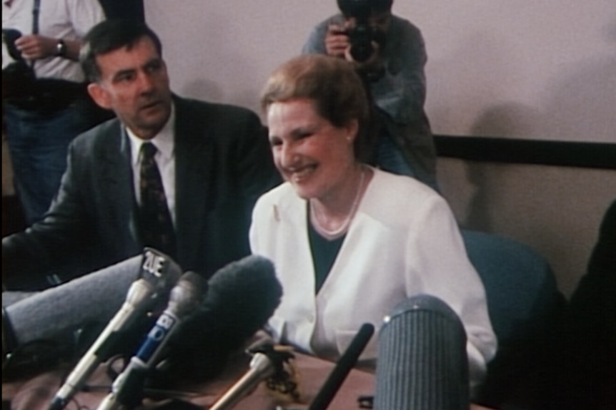 A woman in a white suit jacket smiles behind a bank of microphones