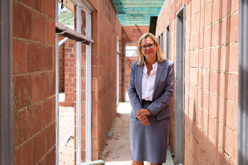 A woman with blonde hair wearing a smart blazer stands in a building site. 