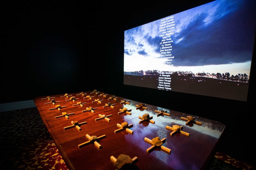 Darkened room with dining table laid with 28 small white crosses with stone on top of each, and video screen hanging over table.