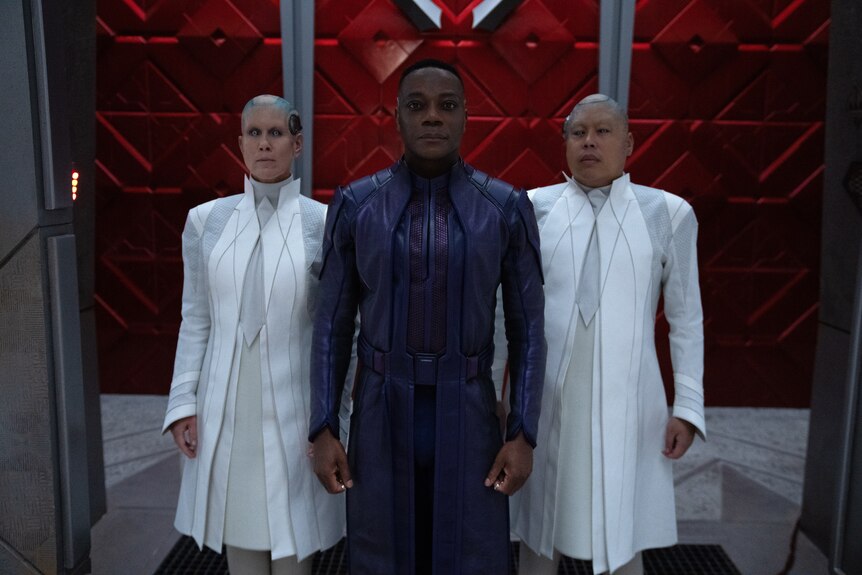 A Black man in a sci-fi black leather suit, flanked by two shaved-headed people in white.