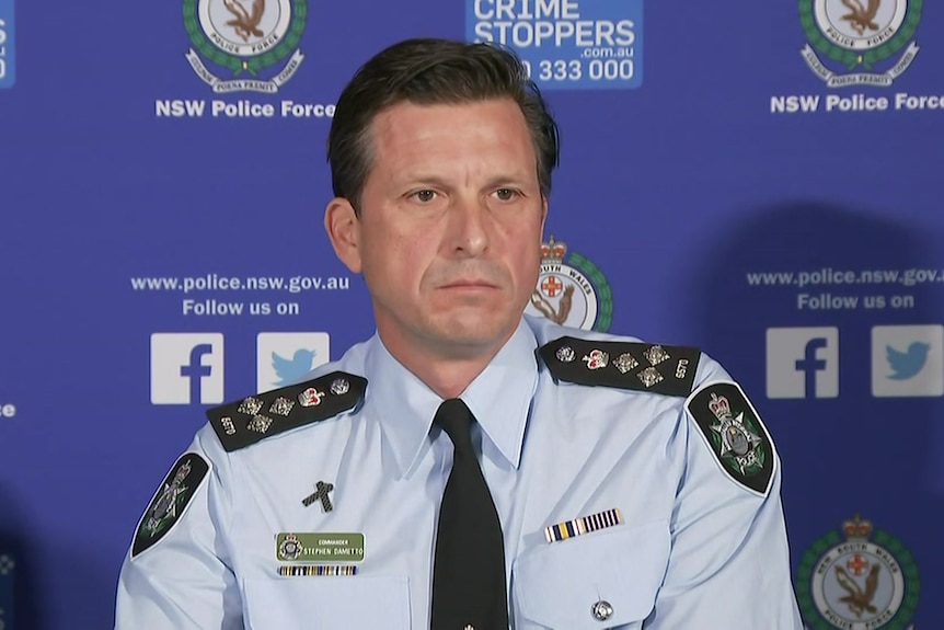 A male police officer in uniform looks serious in front of a police banner at a press conference.