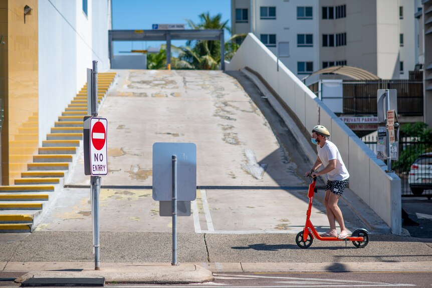 A photo of a man riding an orange scooter in Darwin while wearing a face mask. He is wearing a white T-shirt and blue shorts.