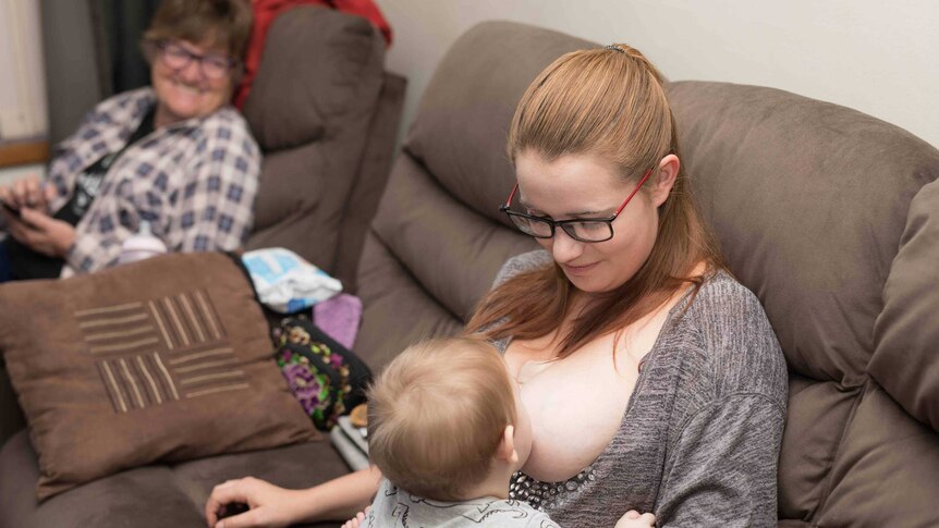 Young woman wearing glasses breastfeeding a baby with an older woman in the background.