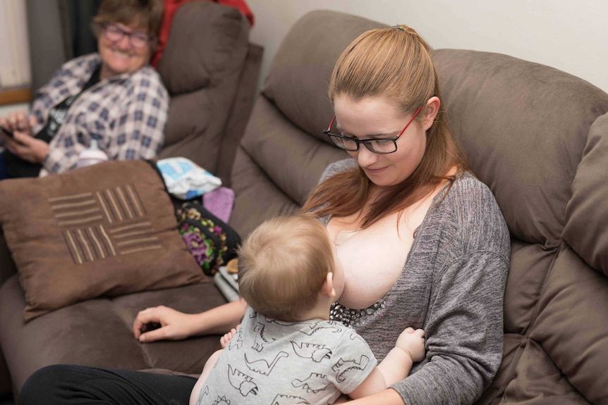 Young woman wearing glasses breastfeeding a baby with an older woman in the background.