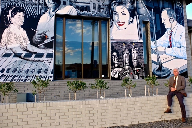 An older man in a suit sits on a ledge next to a mural featuring an image of him broadcasting the news when he was younger.