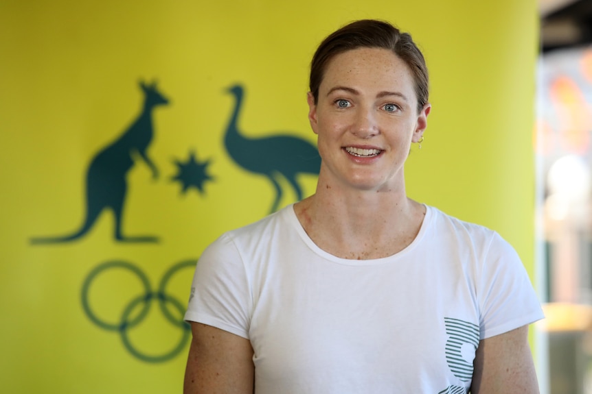 Australian swimmer Cate Campbell, wearing a plain white t-shirt, stands in front of the Australian Olympic coat of arms.