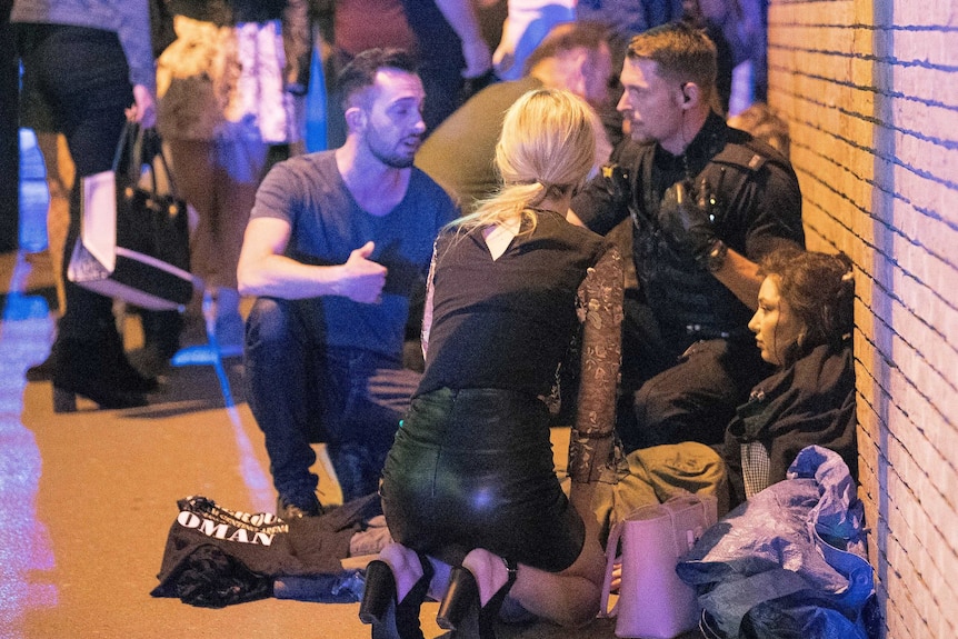 People huddle together around a girl sitting against a brick wall, covered in others' coats.