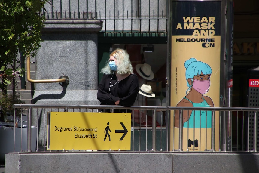 A blonde woman in a face mask next to a sign saying 'wear a mask and Melbourne on'.