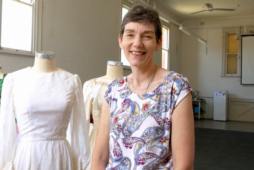 A woman with short brown hair smiles. She is wearing a purple paisley top and there are wedding dresses in background