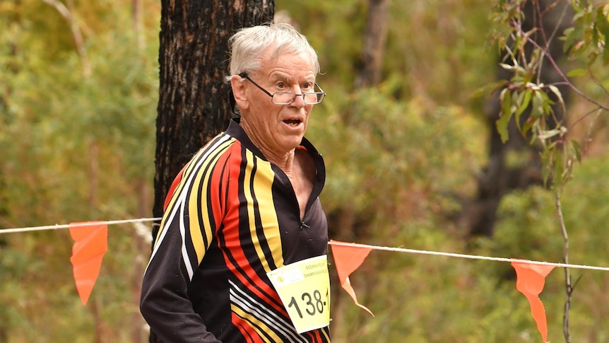 Elderly man in glasses, running in bush, tree behind him, orange bunting