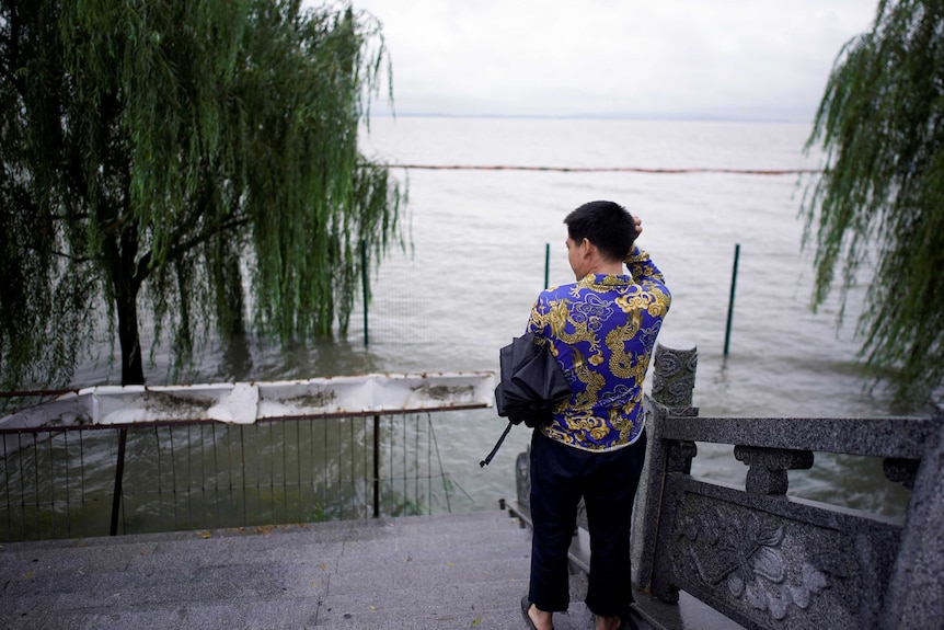 A man in a purple shirt with a dragon pattern stands on the banks of a flooding lake.