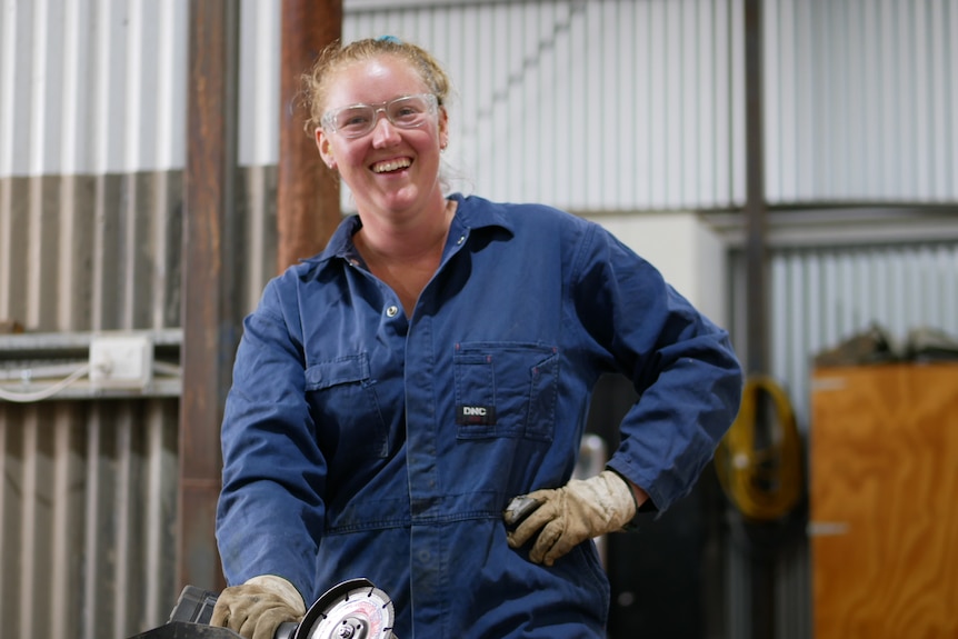 A woman with safety goggles and overalls laughs while looking at the camera