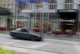 Blurry vision of a dark green sedan driving along the tram tracks in front of shops in Bourke Street Mall.