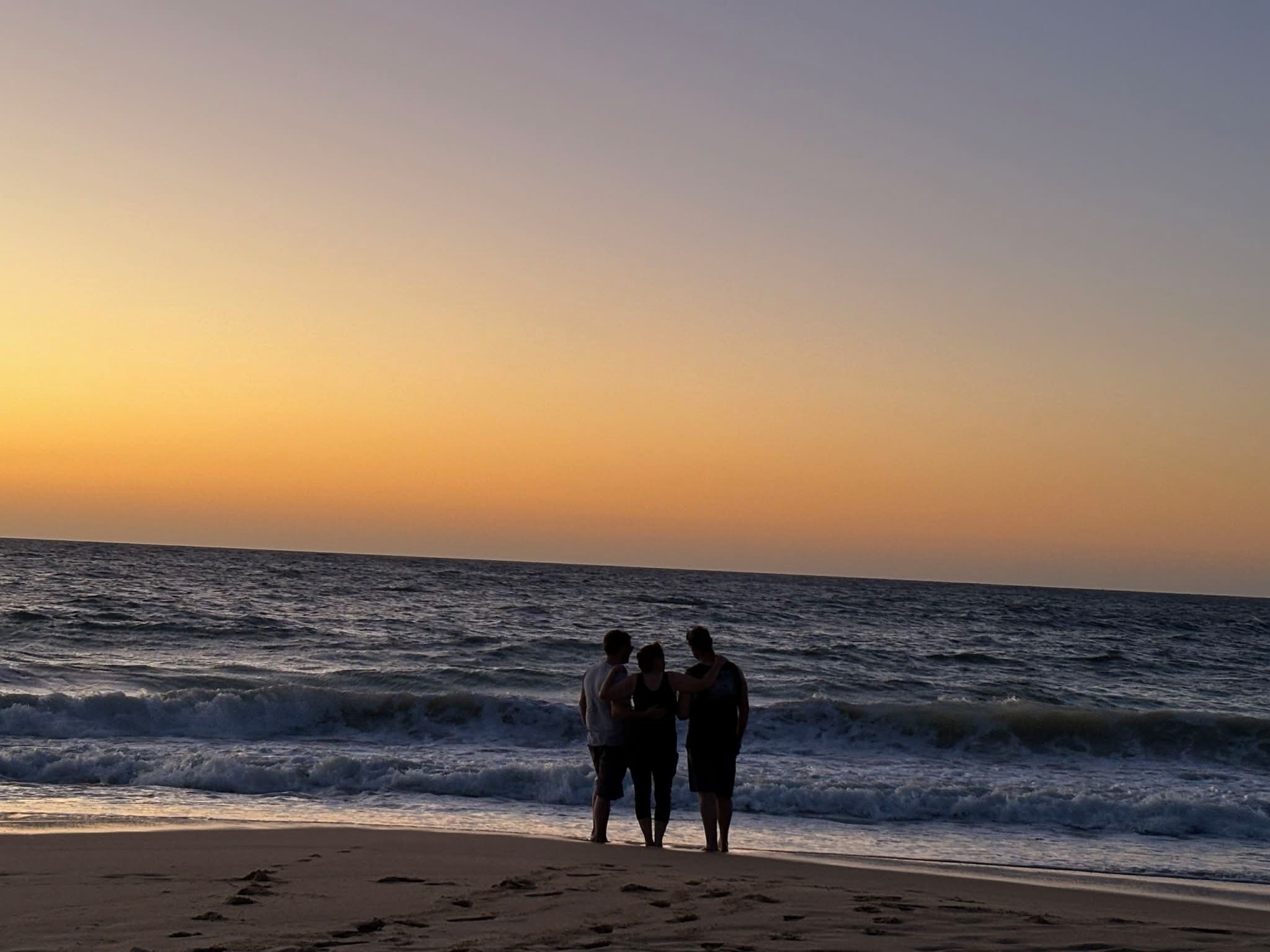 Três pessoas estão em frente ao oceano ao pôr do sol.