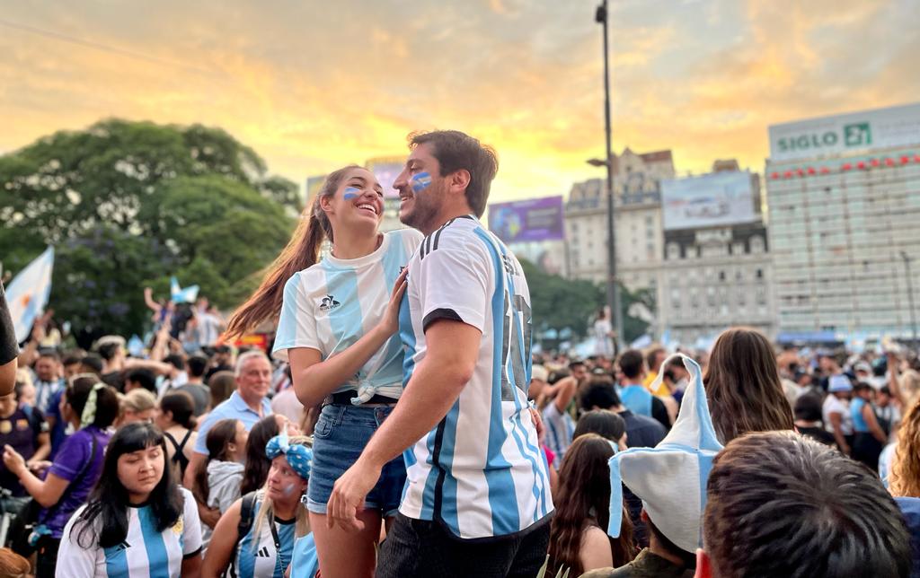 A young man and woman wearing Argentina jerseys and face paint dance in a crowd outdoors.
