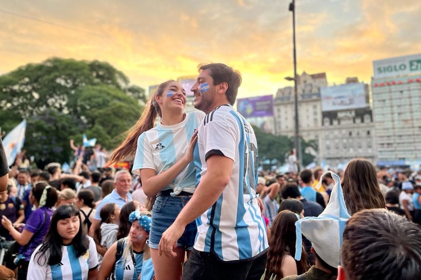 A young man and woman wearing Argentina jerseys and face paint dance in a crowd outdoors.