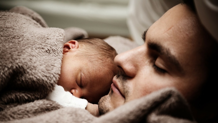 A newborn baby lies on fathers chest, both have their eyes closed.