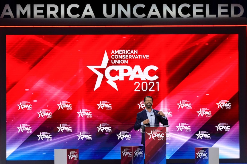 A bearded middle aged man in suit speaks on stage with 'American Uncanceled' sign behind him.