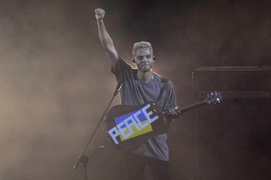 Malik Harris raises his fist in the air and shows a Ukrainian flag with the words peace written on it on the back of his guitar.