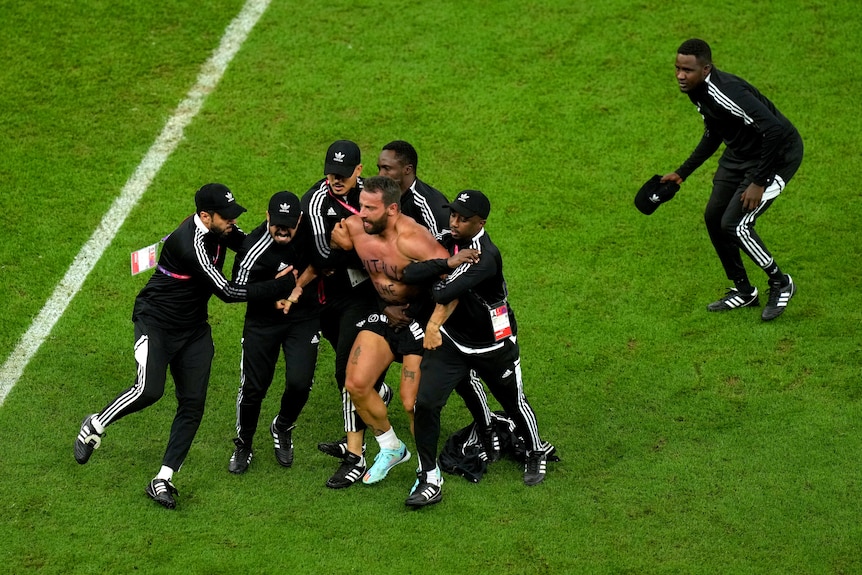 A shirtless man is carried from the field by five security guards in black