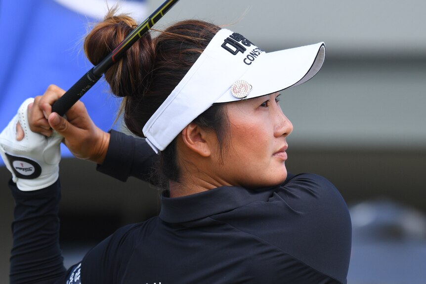 A woman wearing a hat looks into the distance after she has swung a golf club