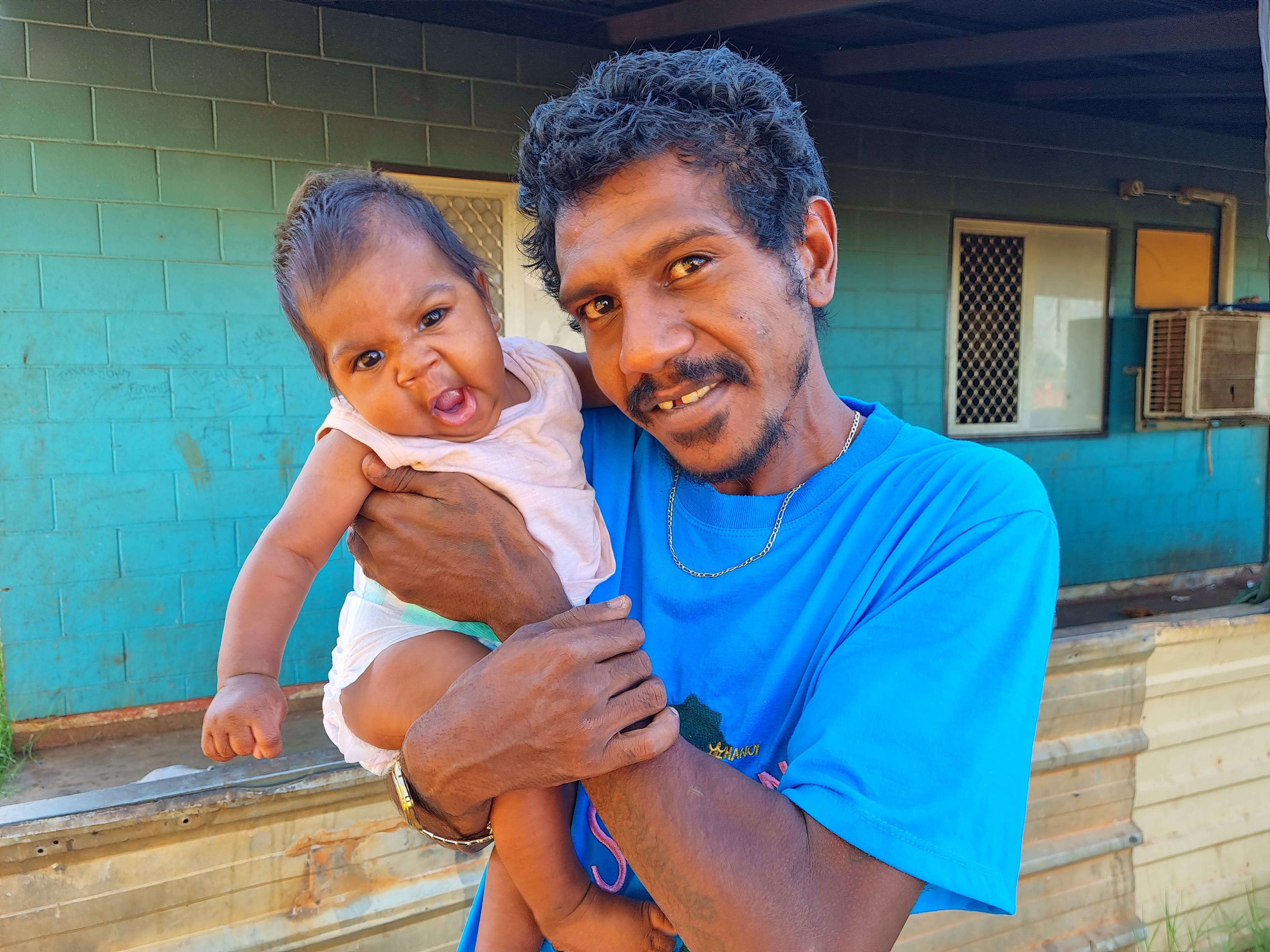 A man wearing a blue shirt with a smiling baby looks at the camera.