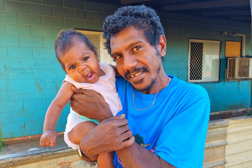 A man wearing a blue shirt with a smiling baby looks at the camera.