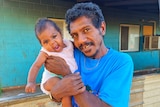 A man wearing a blue shirt with a smiling baby looks at the camera.