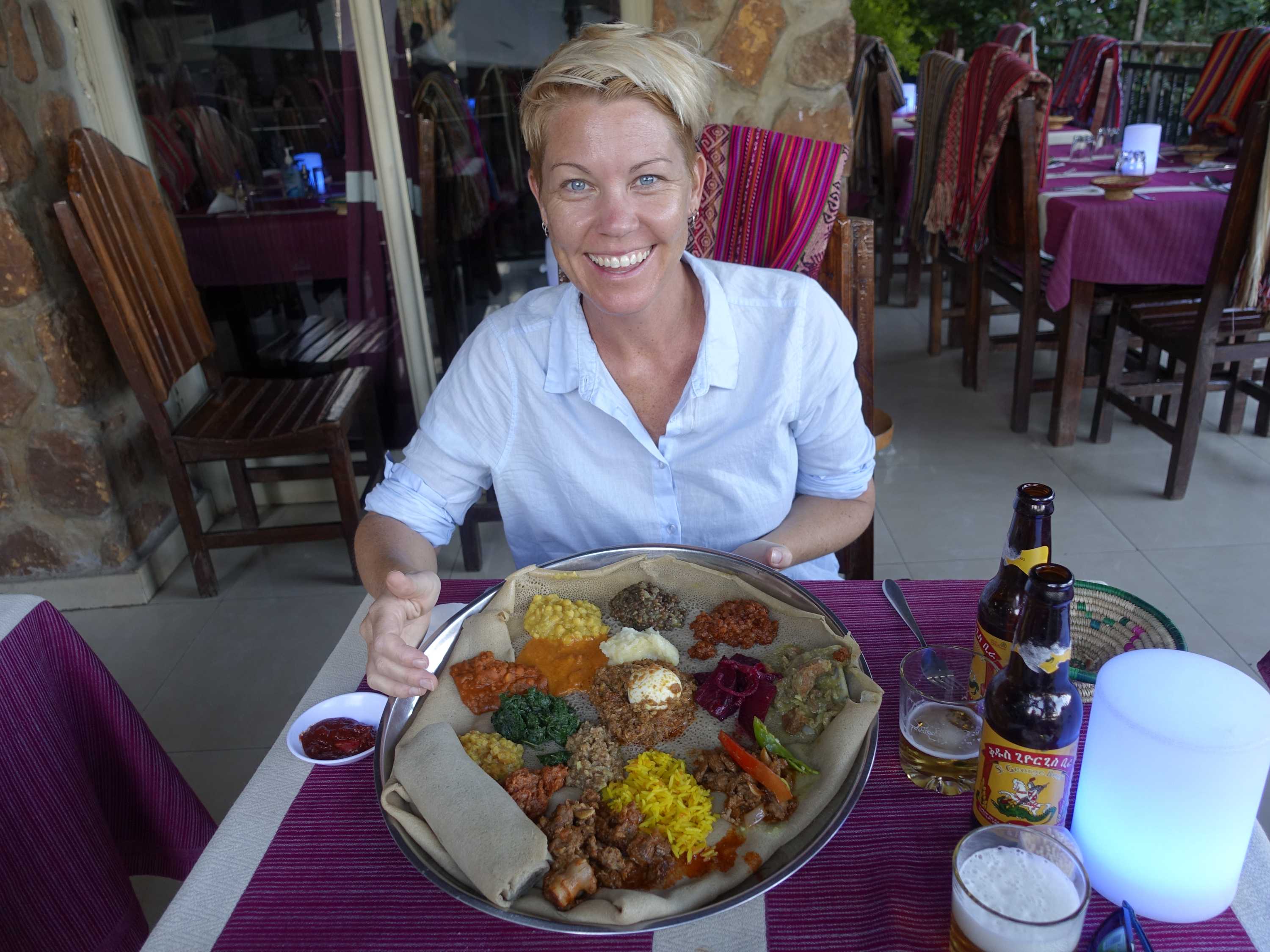 A blonde woman wearing a white shirt sits at a table with a tray of Ethiopian food in front of her