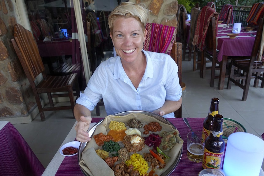 O femeie blondă într-o cămașă albă stă la o masă cu o tavă cu mâncare etiopienă în fața ei