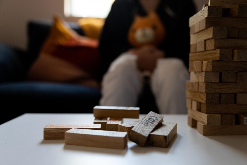 A teenage girl sits on a couch hugging a cat plushie in front of a table with wooden blocks labelled with various words.