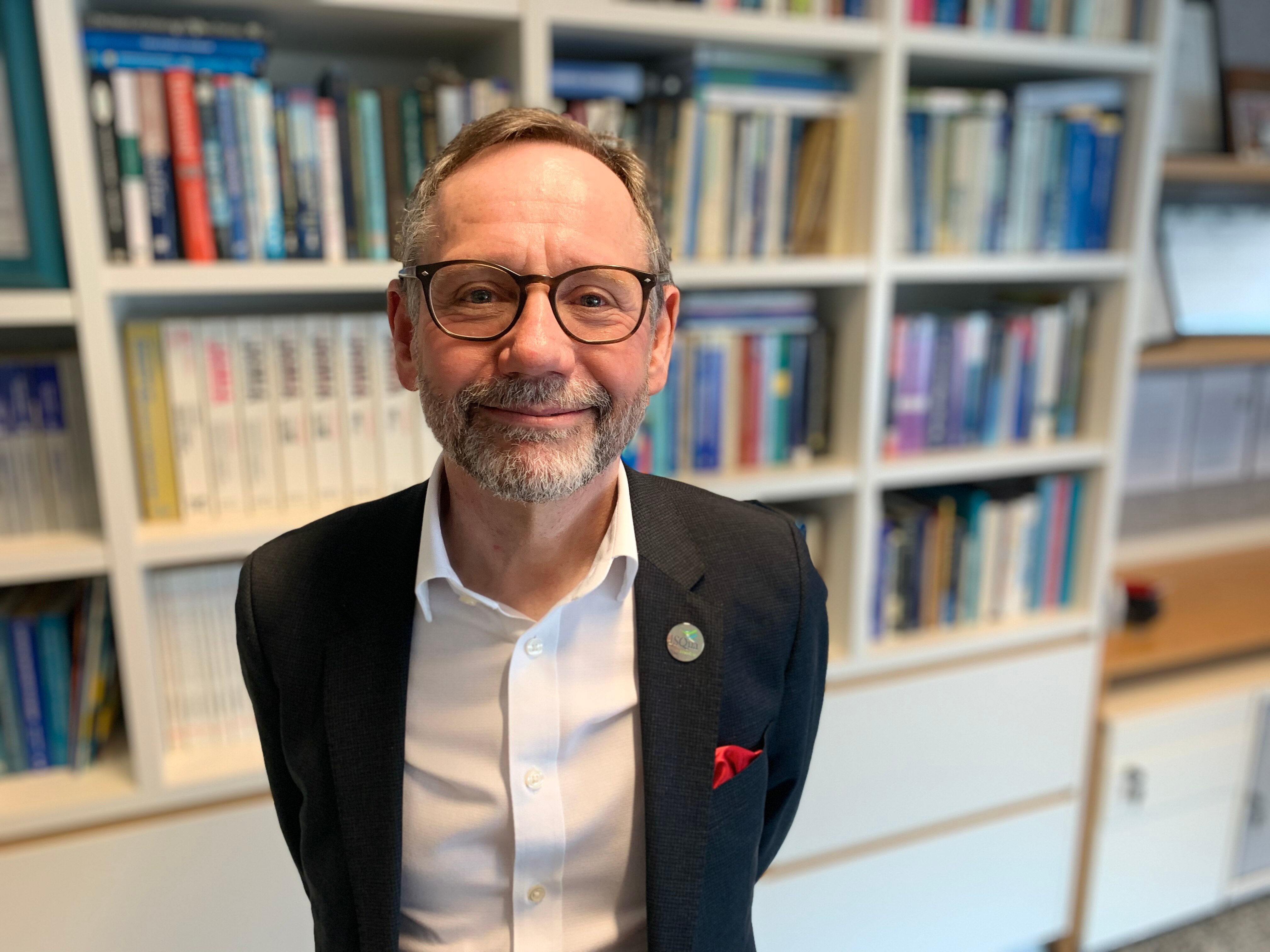 Man wearing a white shirt with black suit jacket and glasses standing in front of a bookcase.
