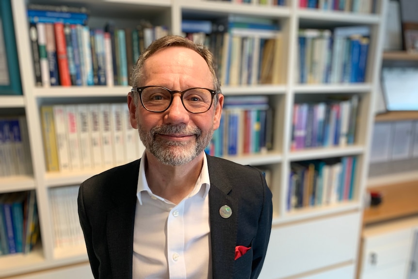 Man wearing a white shirt with black suit jacket and glasses standing in front of a bookcase.